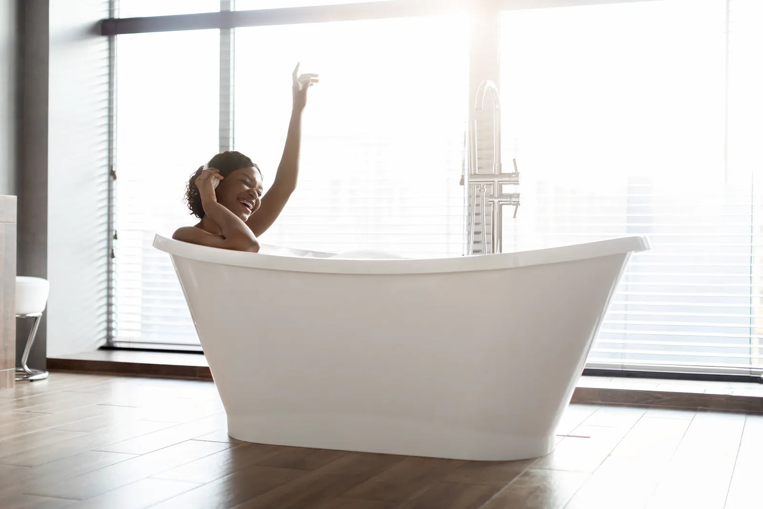 A woman relaxes in a 59-inch white acrylic soaking tub in a bright modern bathroom with large windows.