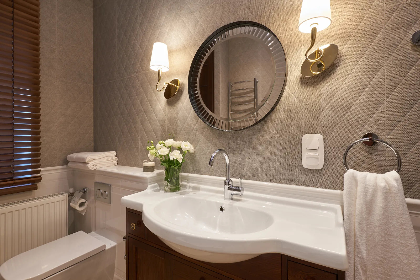Round decorative bathroom mirror with brass sconces above a classic white vanity in a neutral-toned bathroom.