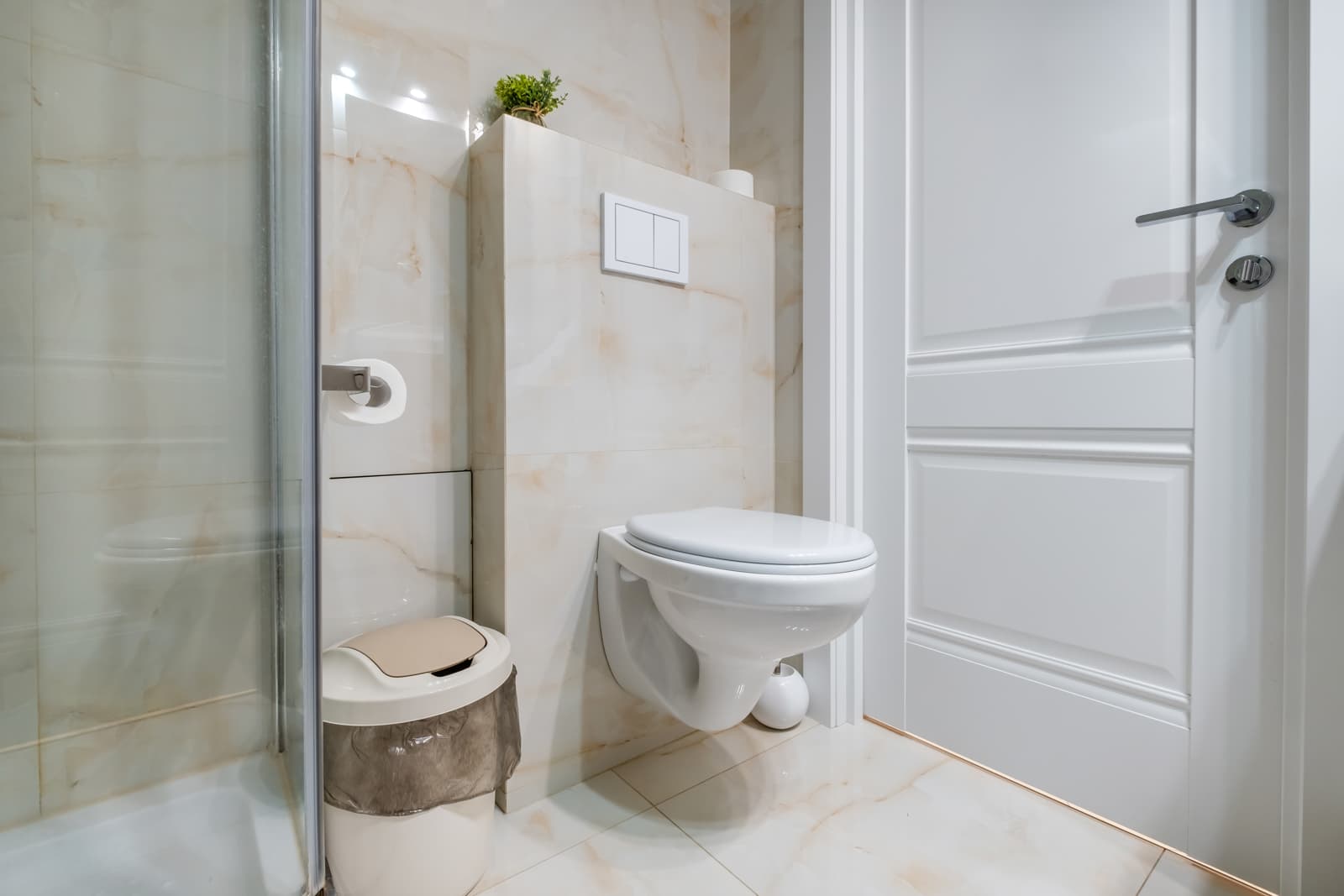 This bright contemporary bathroom features a framed glass shower enclosure beside a wall-mounted white toilet and light marble tile walls.