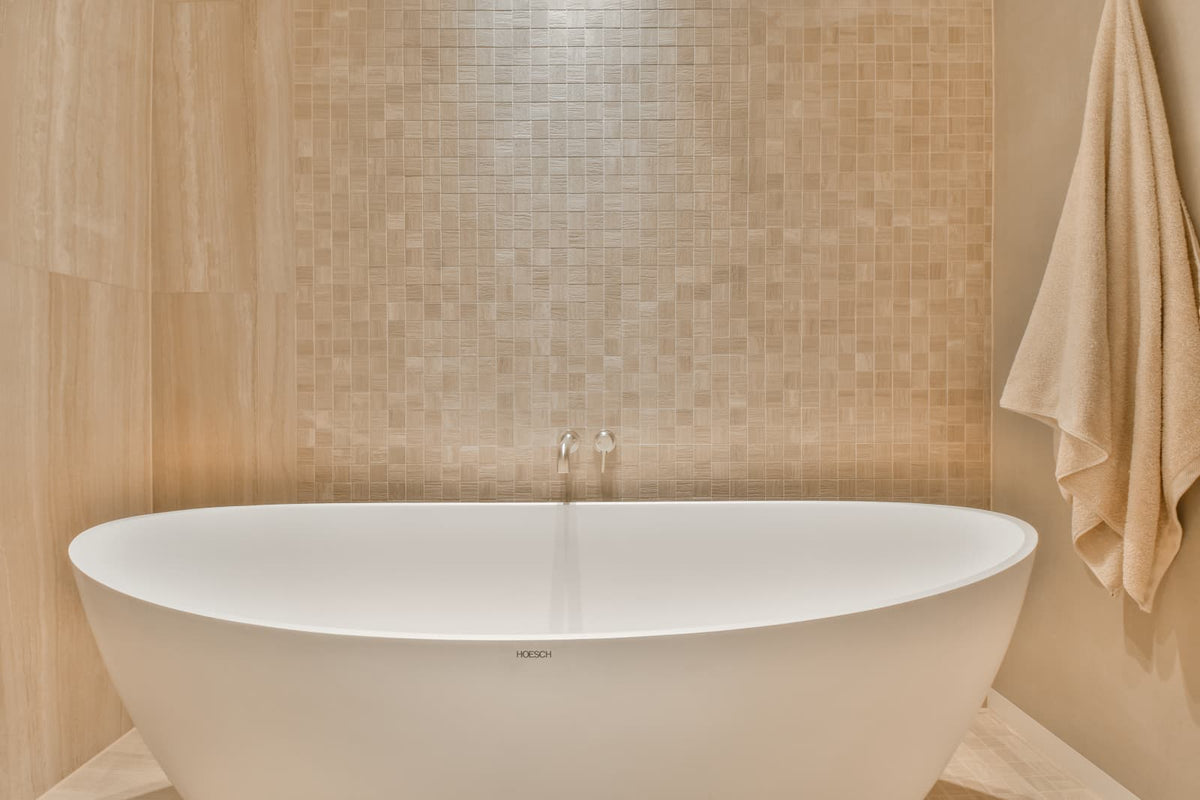 White freestanding oval tub beneath a wall-mounted faucet in a warm beige tiled bathroom.