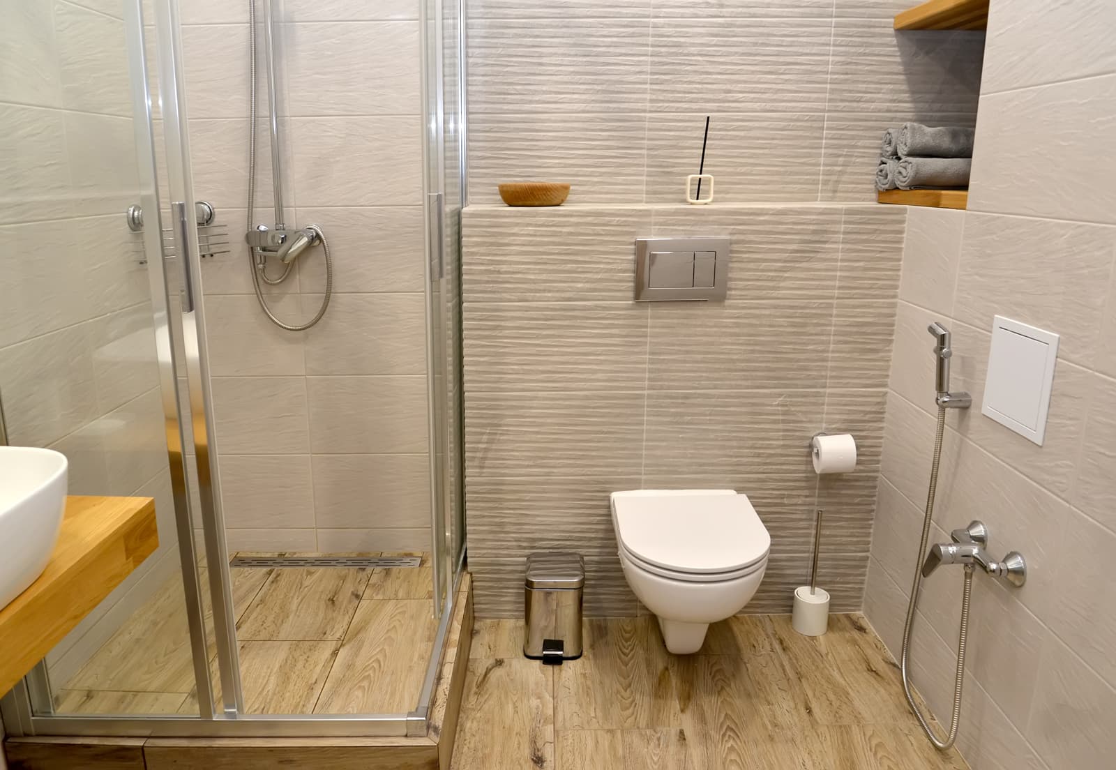 This contemporary neutral bathroom includes a glass shower partition, wall hung toilet and wooden countertop with light wood-look flooring.