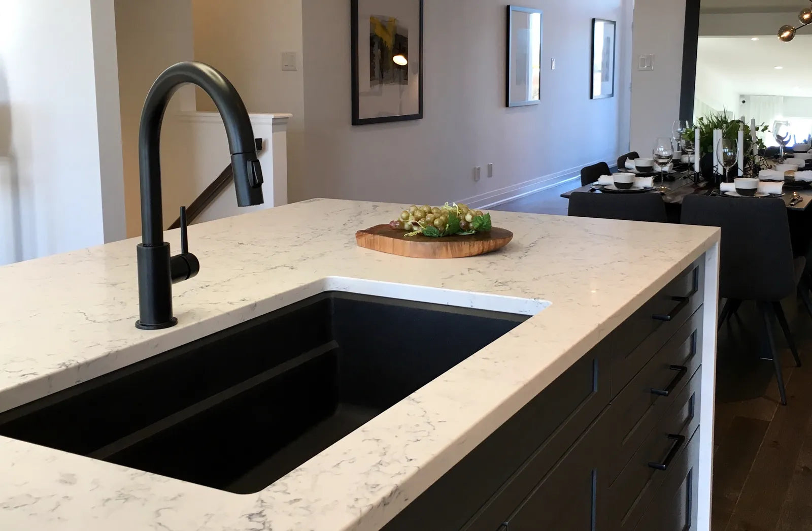 A large black undermount sink in a white quartz kitchen island, with a black faucet and a wooden cutting board of grapes on the counter.