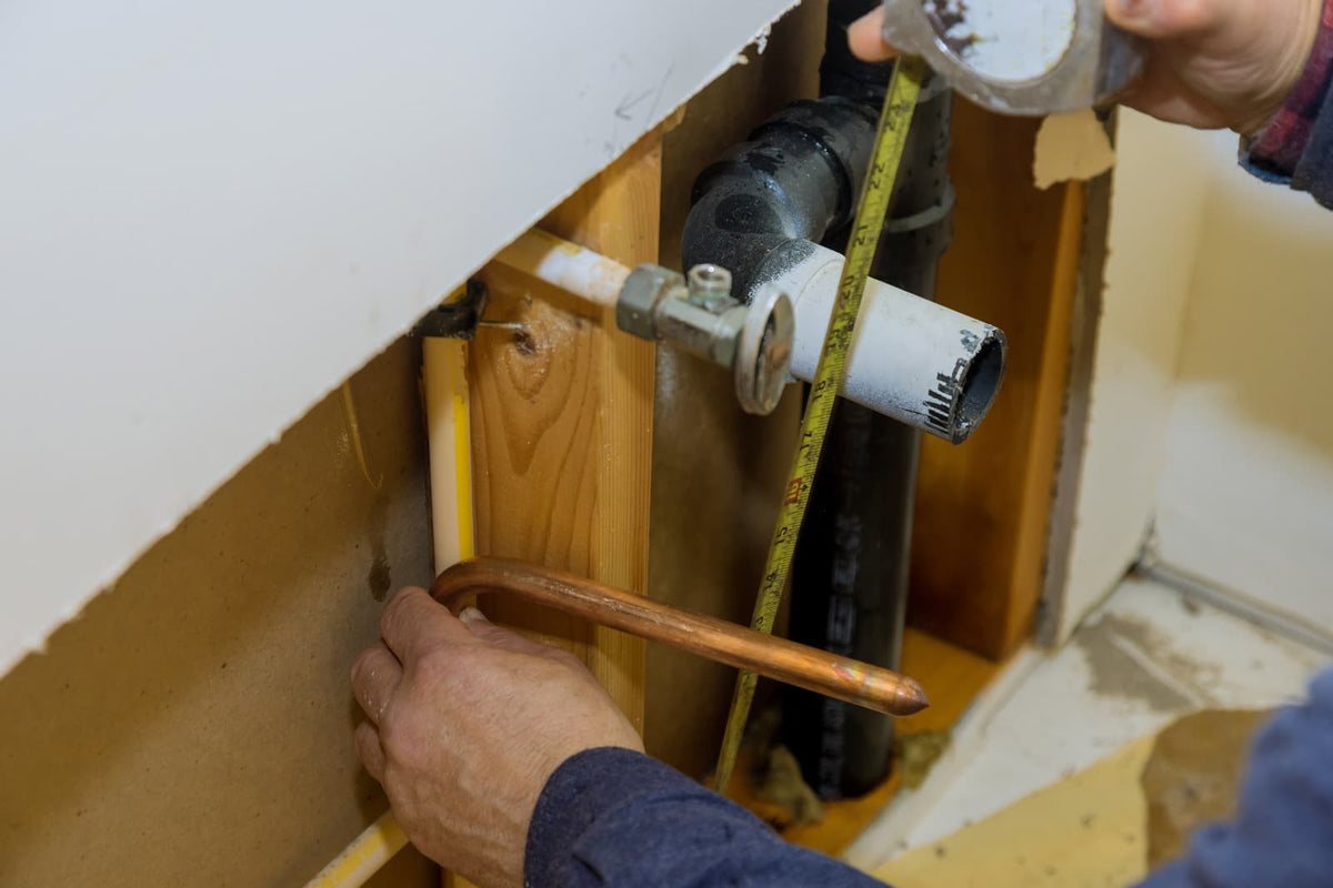 Plumber measures exposed drain and supply pipes beside a bathtub during bathroom renovation.
