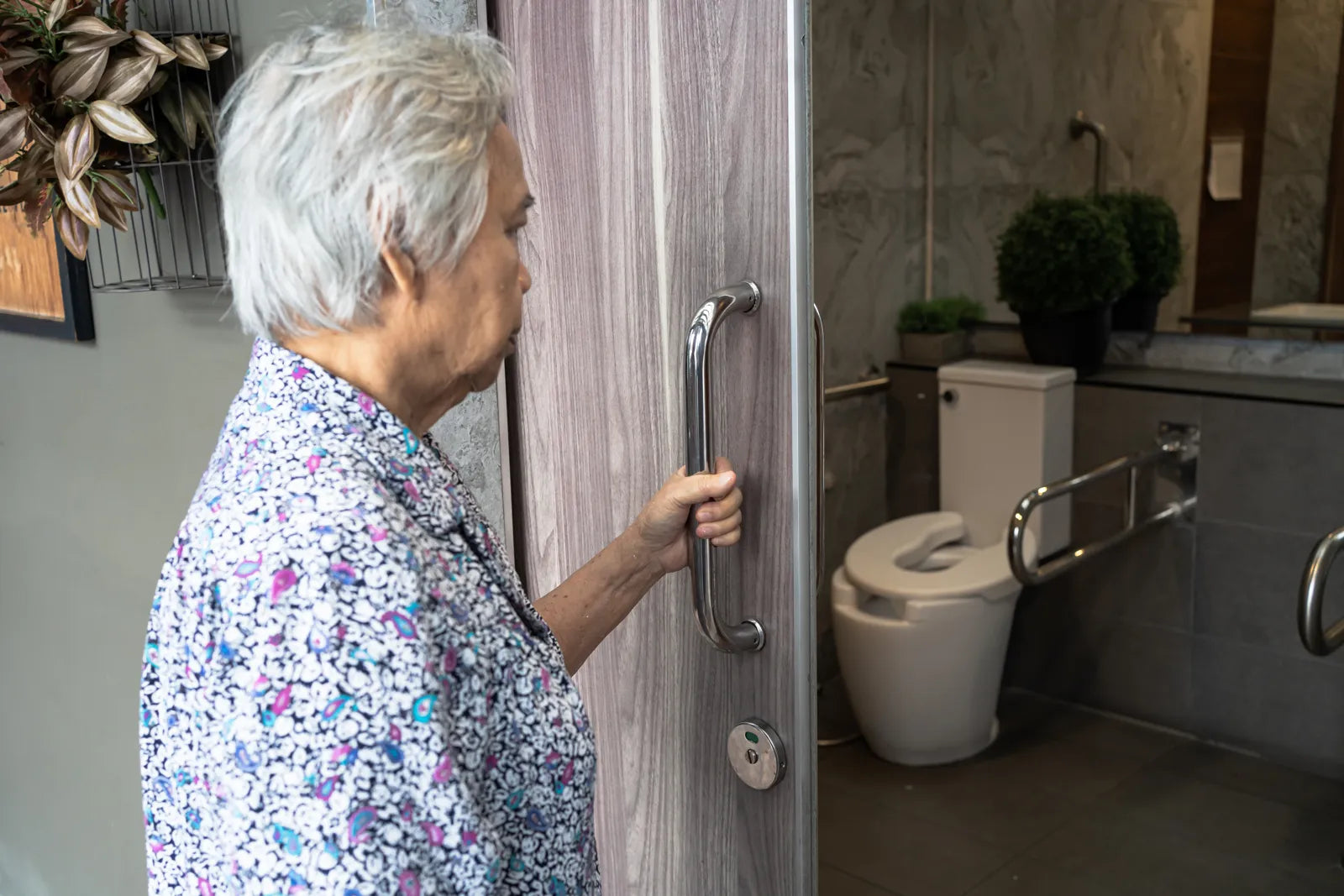 An elderly woman opens the door to an accessible bathroom with grab bars and a comfort-height toilet.