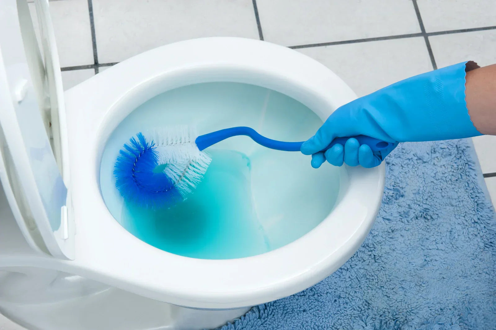 A person wearing blue rubber gloves scrubs a toilet bowl with a blue-and-white brush, performing routine bathroom cleaning to maintain hygiene.