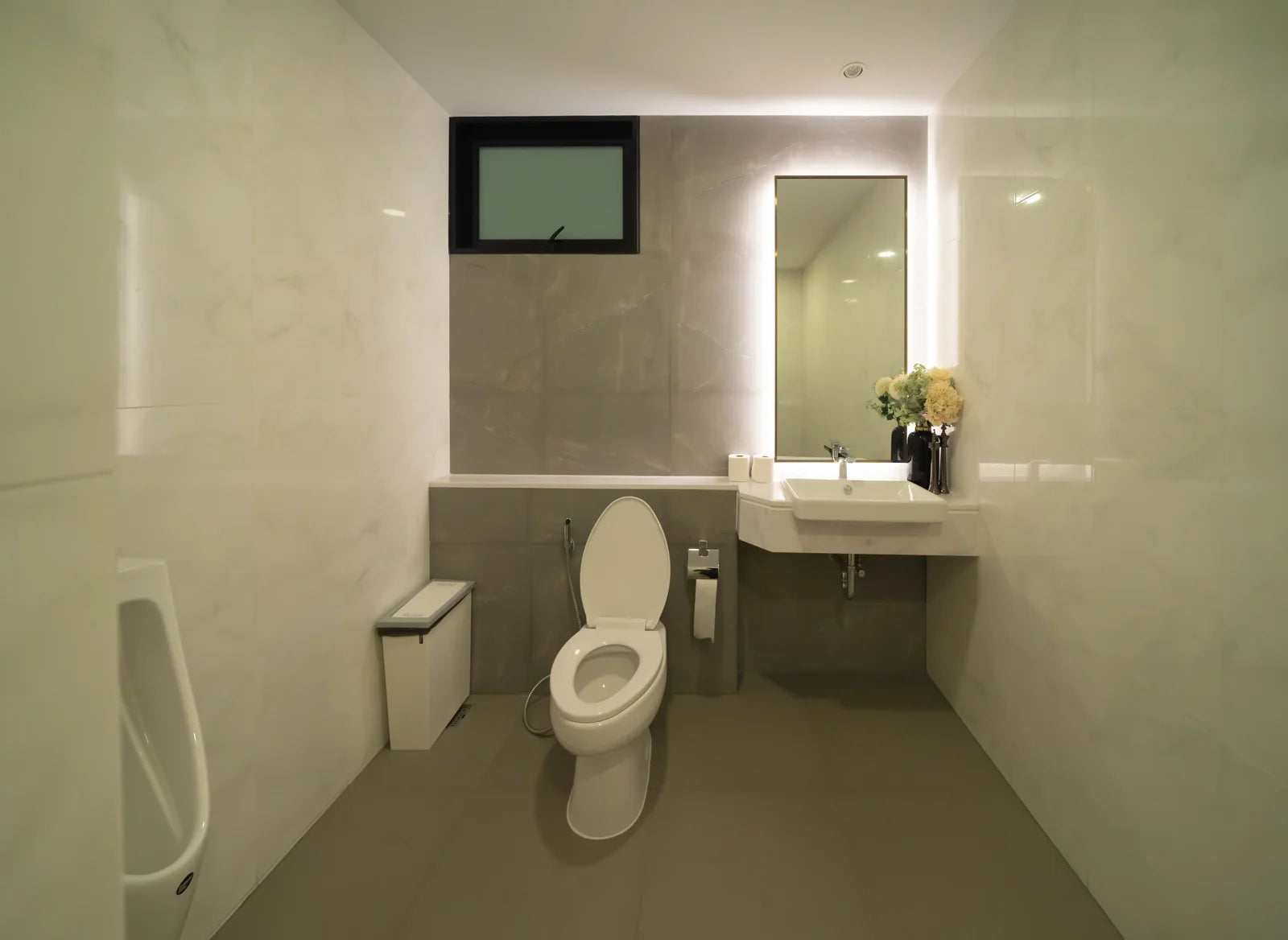 A modern bathroom with a standard toilet, urinal, and wall-mounted sink in neutral tones.