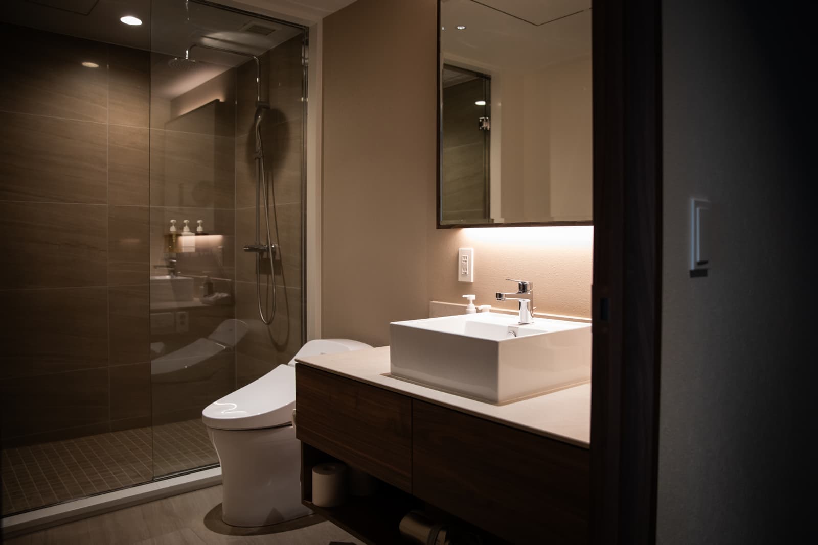 Contemporary warm neutral bathroom featuring a glass shower enclosure, floating vanity, toilet and layered interior lighting.