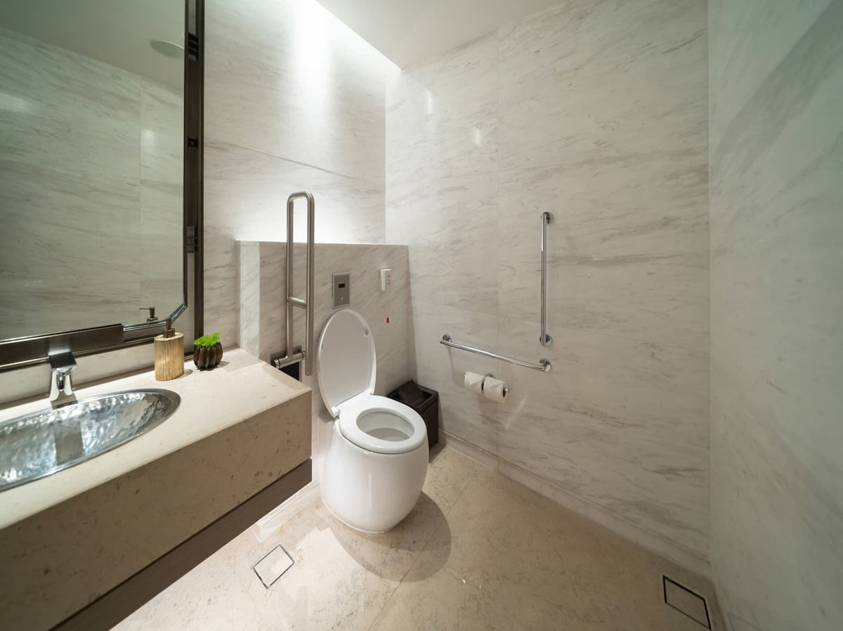 Modern bathroom with an open white toilet, grab bars, stone walls, and a nearby sink.