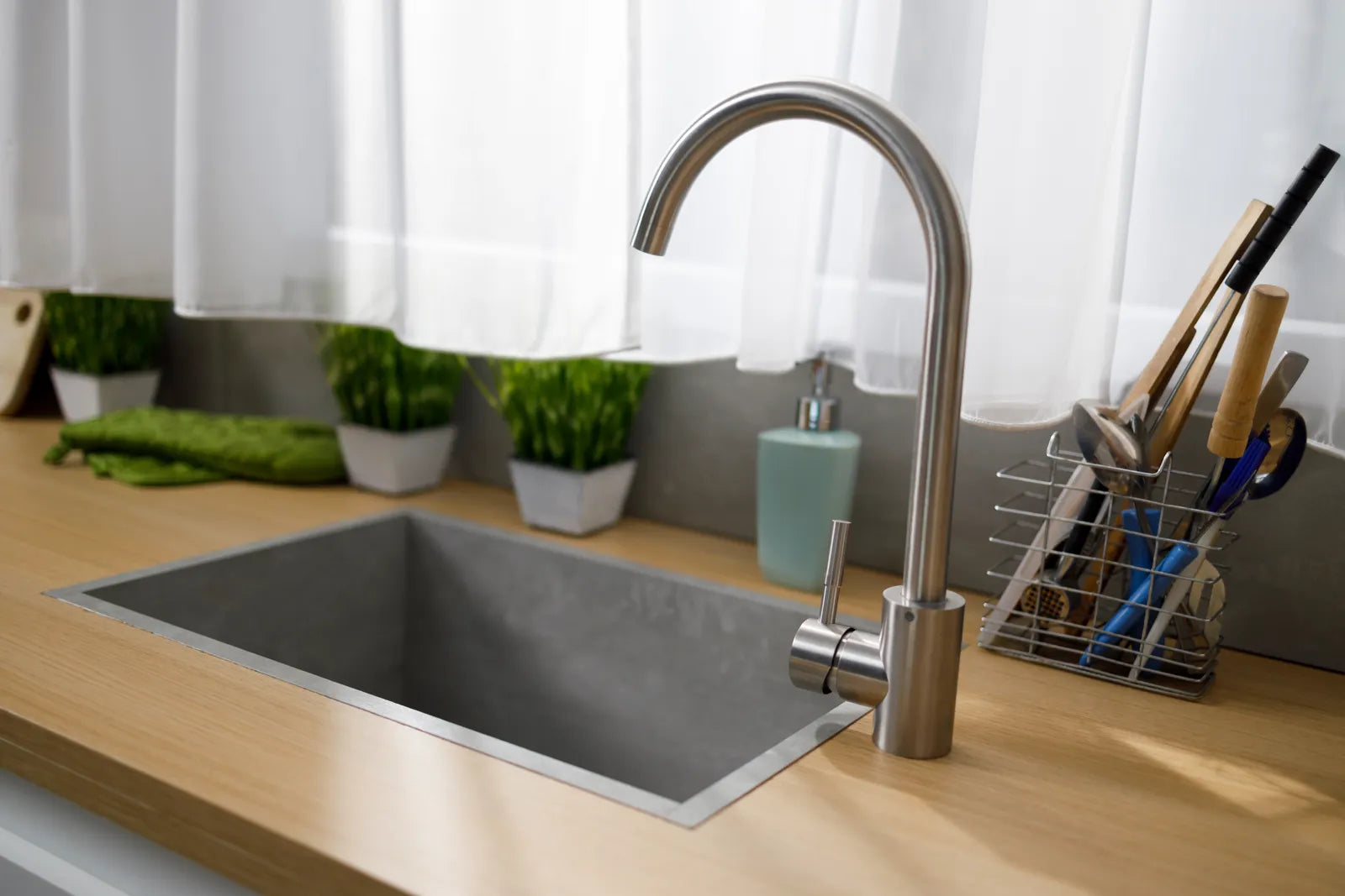 A clean, modern kitchen sink with a stainless steel faucet, wooden countertop, and fresh plants nearby.