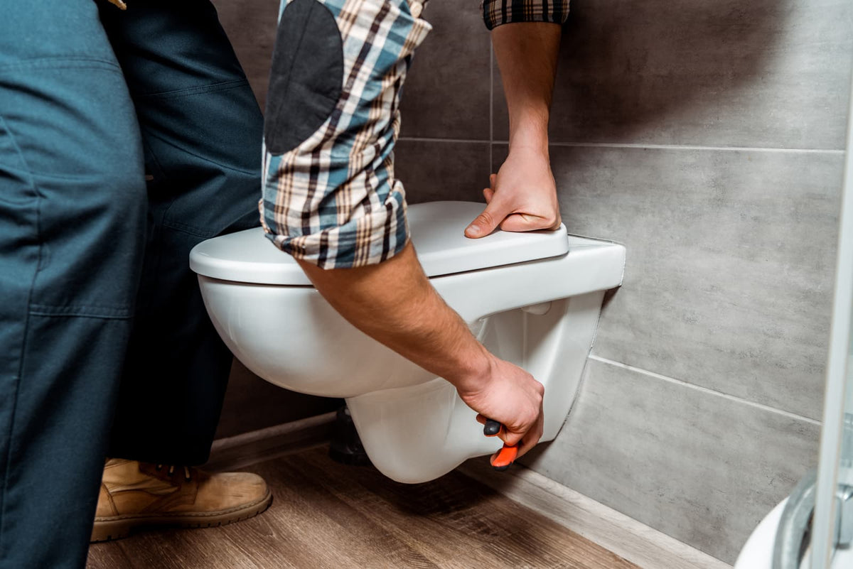 Installer secures a wall-hung toilet bowl against a tiled wall during gasket and mounting service.